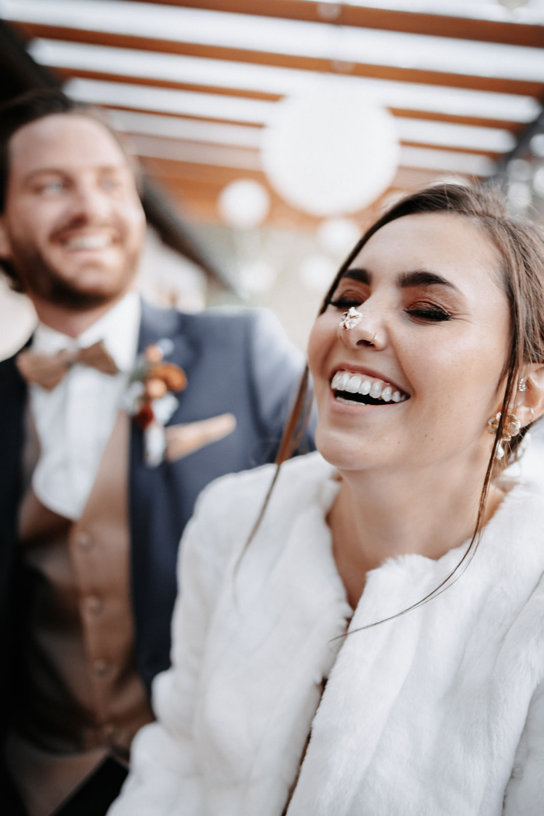 Braut-in-Felljacke-lacht-mit-Sahne-auf-der-Nase-Bräutigam-im-Hintergrund-glückliches-Paar-während-der-Hochzeitsplanung-entspannt-heiraten-mit-Weddingplanner-Augsburg-Nürnberg-Ingolstadt