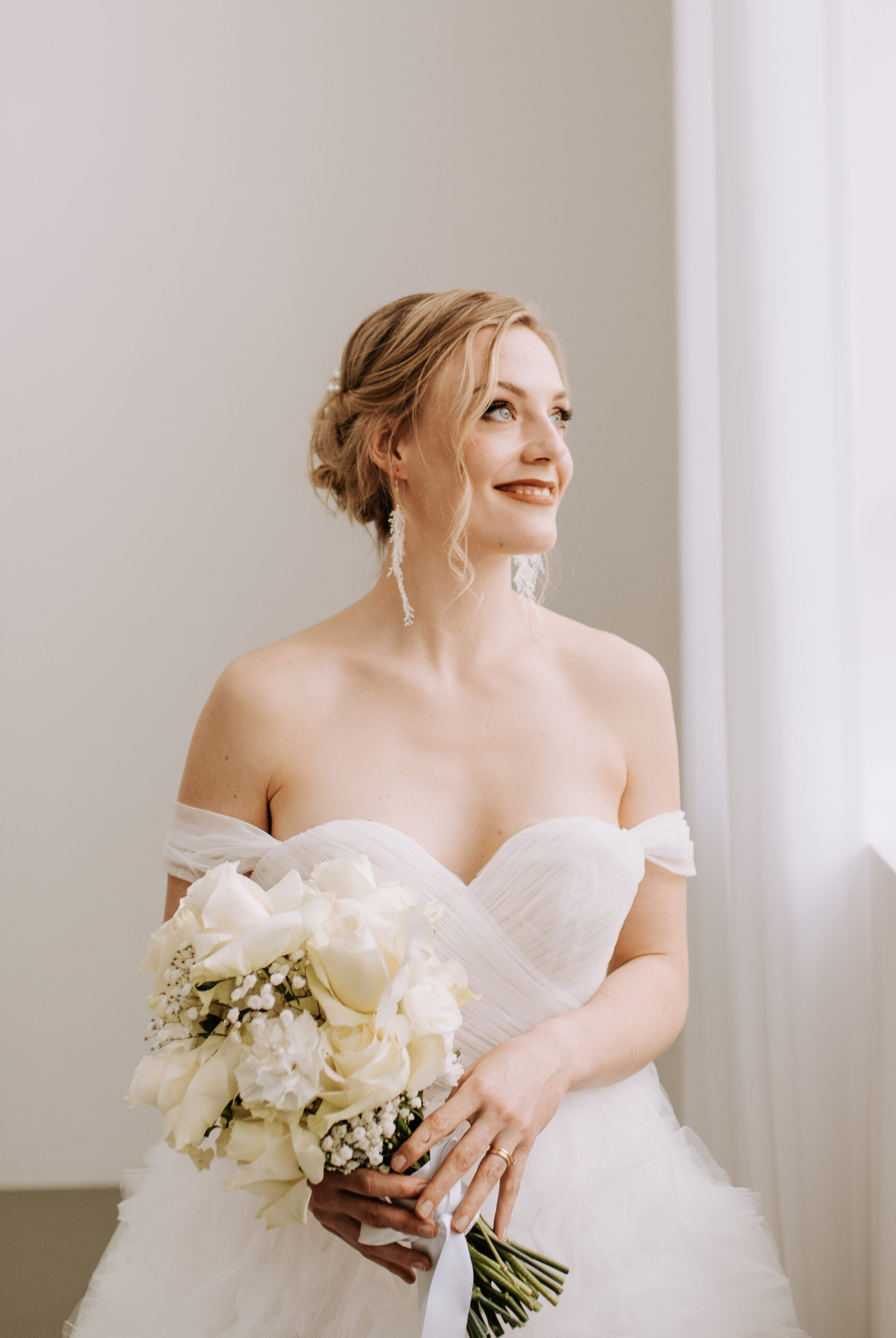 blonde-Braut-mit-weißem-Brautstrauß-und-Prinzessinnenkleid-blickt-glücklich-aus-dem-Fenster-zufriedene-Braut-Heiraten-mit-Hochzeitsplanerin-Nürnberg-Augsburg-Ingolstadt