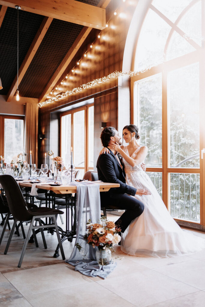 Brautpaar-am-Hochzeitstag-vor-großem-Fenster-in-lichtdurchfluteter-Location-an-Hochzeitstafel-Hochzeitsdeko-Augbsurg-Ingolstadt